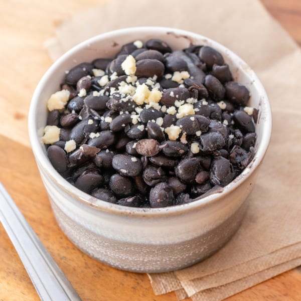 A bowl of black beans topped with crumbled white cheese and a spoon on a wooden table.