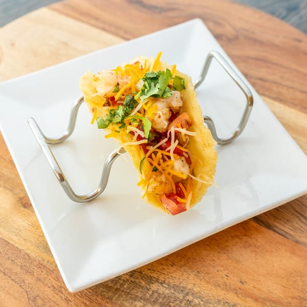 A shrimp taco in a hard shell, topped with cheese, tomatoes, and cilantro, in a metal holder.