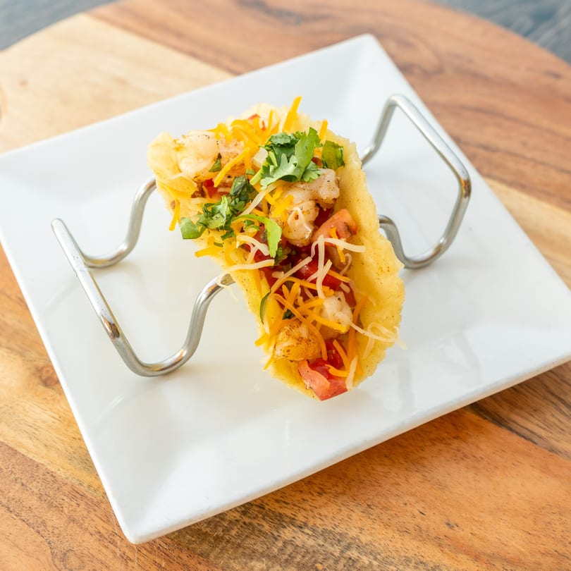 A shrimp taco in a hard shell, topped with cheese, tomatoes, and cilantro, in a metal holder.
