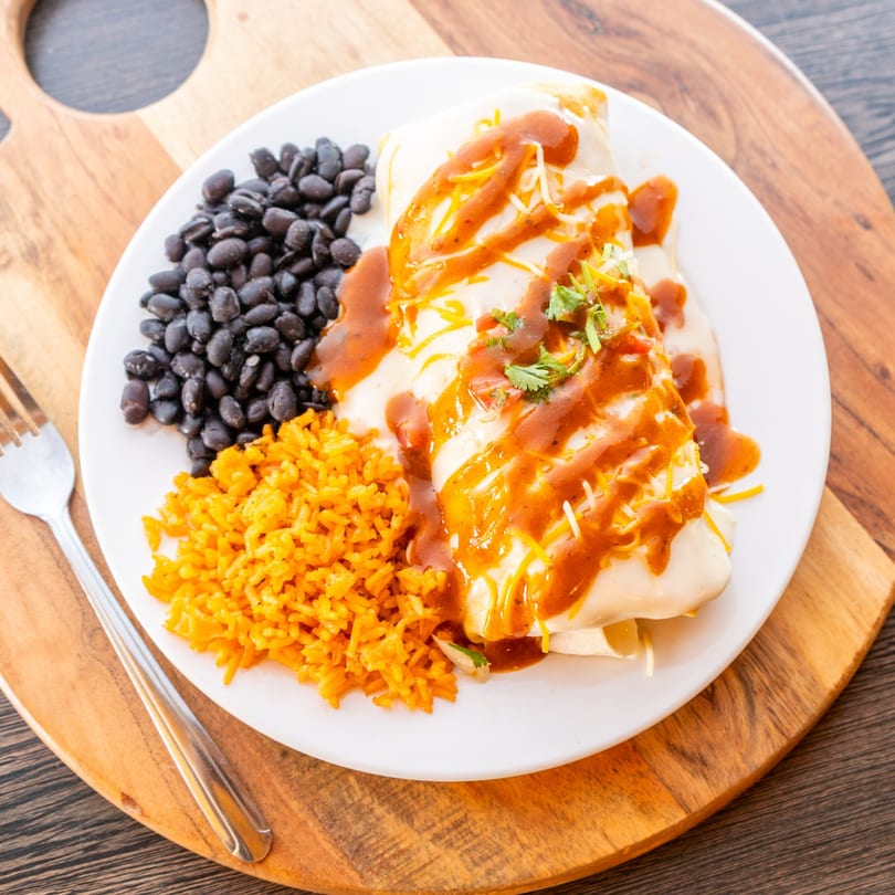 A burrito covered in white and red sauce, with a side of black beans and orange rice.