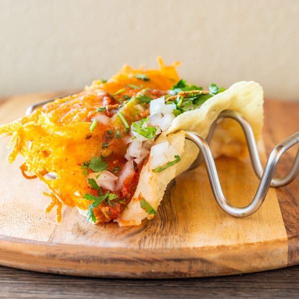 A crispy birria taco with melted cheese, cilantro, and onions in a metal holder on a wooden board.