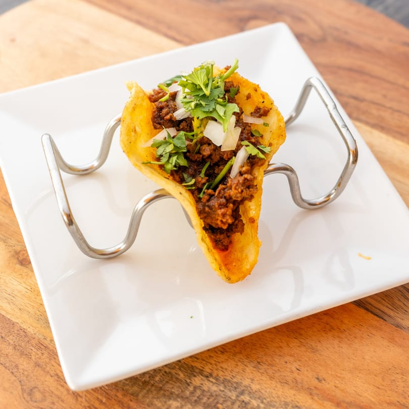 A single taco with ground meat, cilantro, and onions in a crispy shell, held by a metal stand.
